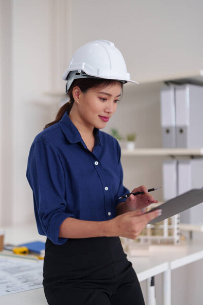 Asian architect woman professionally reviews building plans at an office. Her focus on design innovation ensures project success.