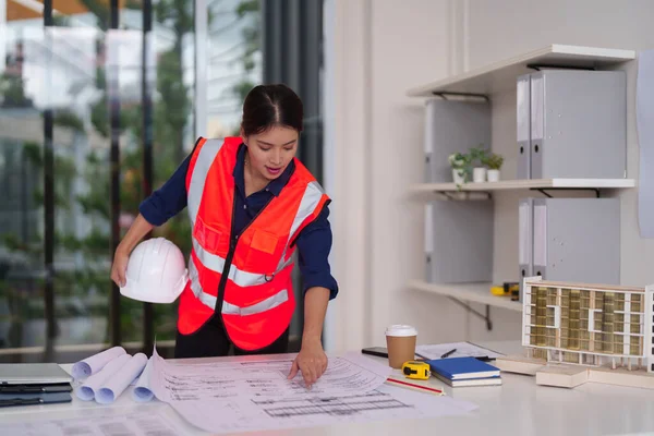 Odaklanmış Asyalı mimar, geleceğin kentsel inşaatını tasarlayan çelik yelek giyen modern bir ofisin ayrıntılı bina planlarını gözden geçiriyor.