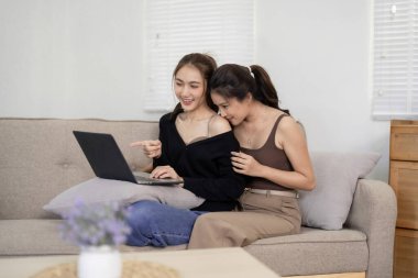 İki Asyalı ortak, dizüstü bilgisayarlarını kullanarak öğleden sonra evlerinin keyfini çıkarıyorlar. Yumuşak bir salonda birbirlerine bağlanarak gülümsüyorlar..