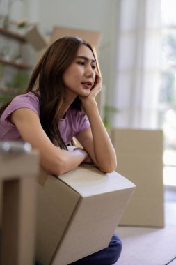 Genç Asyalı kadın, etrafı karton kutularla çevrili yerde oturuyor. Yeni başlangıç ve yer değiştirme sürecini düşünürken çok düşünceli görünüyor.