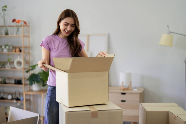 Веселая взрослая азиатка распаковывает вещи в новой квартире. Она улыбается, находя предметы в своем современном доме. Радость переселения.