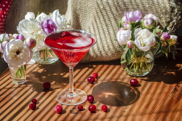 Bodegón rústico solemne con vaso de bebida roja con bayas de otoño y hielo rodeado de flores blancas sobre una mesa de madera a la sombra diagonal de las persianas en un día soleado. Hermosa refracción de luz —  Fotos de Stock