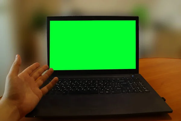 Laptop on a table with a green screen. A man has problems with a computer and shows the screen with his hand. First-person shot.