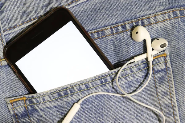 Template for audio and video content. Phone with a white screen and headphones in a jeans pocket. Bright photo to insert your image.
