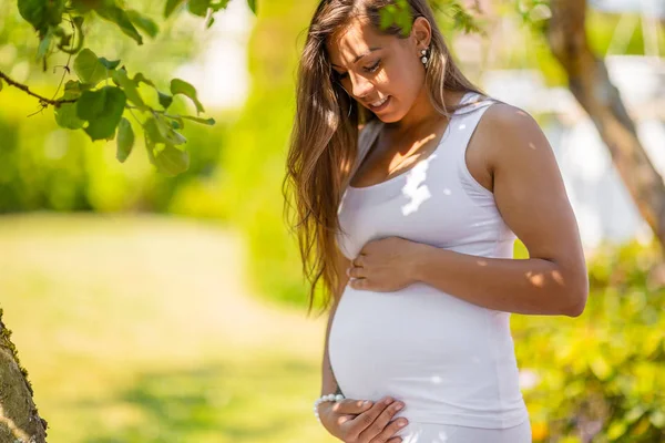 Mosolygó terhes nő áll a kertben kéz a hason — Stock Fotó