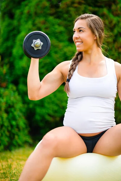 Mulher grávida sorrindo ao levantar halteres na bola de ioga — Fotografia de Stock