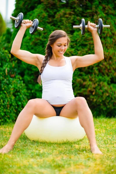 Mujer embarazada segura levantando pesas en la bola de yoga en el parque — Foto de Stock