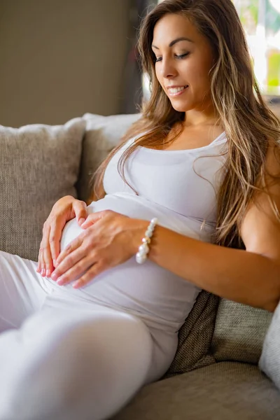 Mulher grávida sorridente no sofá formando uma forma de coração na barriga — Fotografia de Stock