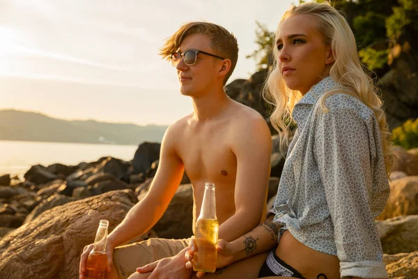 Jeune couple tenant des bouteilles de bière assis sur des rochers à la plage — Photo