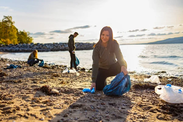 Une jeune femme souriante nettoie la plage avec des bénévoles pendant le coucher du soleil — Photo