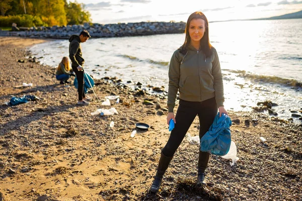 Lächelnde junge Frau putzt Strand mit Freiwilligen bei Sonnenuntergang — Stockfoto