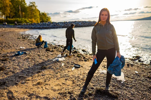 Serious jeune femme nettoyage plage en plastique avec des bénévoles pendant le coucher du soleil — Photo