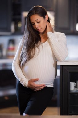 Modern bir mutfak ortamında karnını nazikçe kucaklayan ve derin düşüncelere dalmış hamile bir kadının huzurlu bir görüntüsü. Ortam sıcaklığı ve beklentiyi yansıtıyor. Düşünceli bir durgunluğu yansıtıyor..