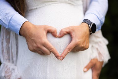 İki elin yakın çekimi hamile bir karın üzerinde kalp şekli oluşturur, aşkı, ilgiyi ve beklentiyi sembolize eder, sakin bir aydınlatma hassas ve içten bir anı vurgular..