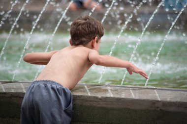 Küçük çocuk yazın sıcaktır ve gömleğini çıkardı ve ısıya daha kolay dayanmak için çeşmeye suyla atlamak istiyor gibi görünüyor. Küçük bir çocuk çeşme suyuna atlar.