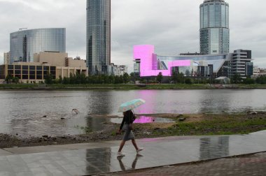 Şehir manzarası, bir nehir, bir gökdelen ve şemsiyeli bir kız. Yağmur yağıyor. Yansımaları ile ıslak kaldırım. Yekaterinburg 2019.