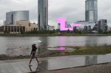 Şehir manzarası, bir nehir, bir gökdelen ve şemsiyeli bir kız. Yağmur yağıyor. Yansımaları ile ıslak kaldırım. Yekaterinburg 2019.