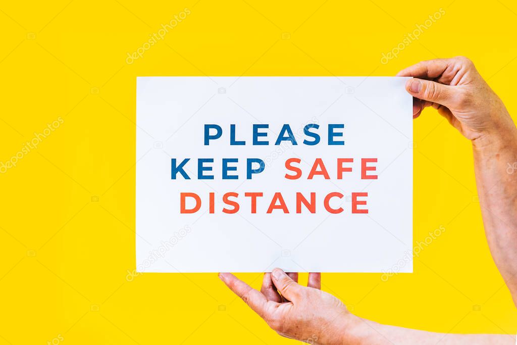 Please keep safe distance. Hands holding a sign for people to respect social distancing measures with an orange background.