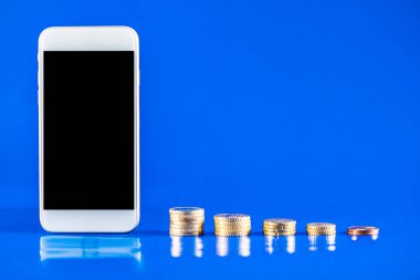 Mavi arka planda bir yığın bozuk para olan cep telefonu. İş büyümesi ve finansal tasarruf kavramı. Kopyalama alanı için siyah ekranlı cep telefonu.