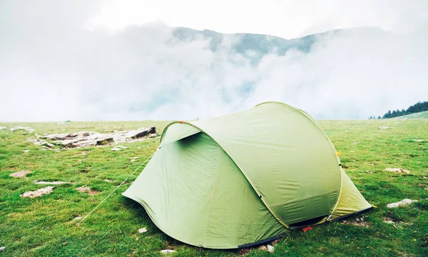 Tent in the wilderness. A single green camping tent in a grassy meadow with fog on the mountain. Trekking, camping, hiking and wild life concept. Real Outdoor Adventure.