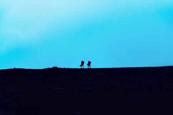 Travelers walk along a trail in the mountains. Silhouettes of two adventure hikers with backpacks walk in the mountains at dusk.