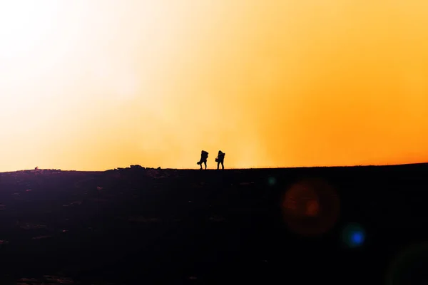 Travelers walk along a trail in the mountains. Silhouettes of two adventure hikers with backpacks walk in the mountains at sunset or sunrise.