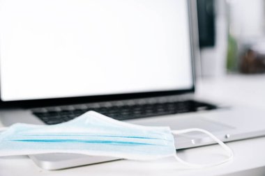 Arka planda odaklanmamış bir dizüstü bilgisayarın üstünde tıbbi yüz maskesi. Coronavirus covid 19 vakalarında evde telgraf çekiliyor..