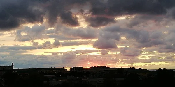 Gloomy autumn clouds over dark silhouettes of urban houses on a sunset background. Black clouds in the foreground are replaced by golden skylight and sky. A striking contrast of light and darkness. Bright picturesque alarming exciting background.