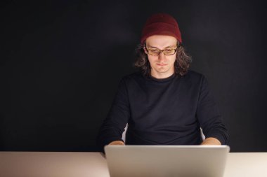 Designer or programmer in the workplace. young man in a hat and glasses sitting at a laptop. clipart