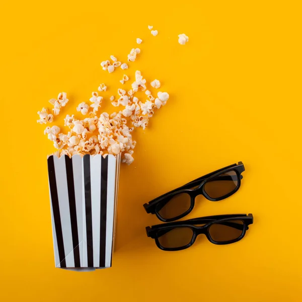 Two black 3d glasses lie next to a black and white papper striped box of popcorn, scattered on a yellow background. — Stock Photo, Image