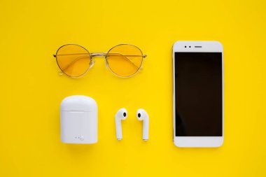 Pembe şeffaf güneş gözlüğü, kablosuz kulaklık ve akıllı telefon parlak sarı bir arka plan üzerinde yatıyor.