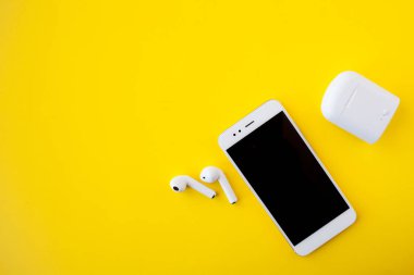 Beyaz kablosuz kulaklık ve akıllı telefon parlak sarı bir arka planda duruyor. Kulaklıklar şarj edilmiyor.