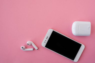 Beyaz kablosuz kulaklıklar ve akıllı telefon parlak pembe bir arka plan üzerinde yatıyor. Kulaklıklar şarj kılıfı dışında.