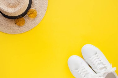 Pembe güneş gözlüğü takan, hasır bir şapkanın önünde sarı arka planda yatan beyaz spor ayakkabılar..