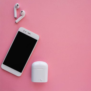 Beyaz kablosuz kulaklıklar ve akıllı telefon parlak pembe bir arka plan üzerinde yatıyor. Kulaklıklar şarj kılıfı dışında.