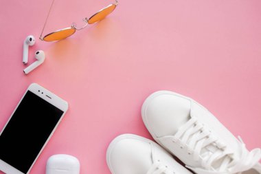 Beyaz spor ayakkabılar, pembe şeffaf güneş gözlükleri, kablosuz kulaklıklar ve akıllı telefonlar pembe arka planda yatıyor..