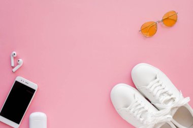Beyaz spor ayakkabılar, pembe şeffaf güneş gözlükleri, kablosuz kulaklıklar ve akıllı telefonlar pembe arka planda yatıyor..