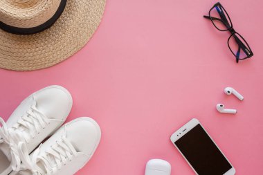 Beyaz spor ayakkabılar, şapka, gözlük, kablosuz kulaklık, akıllı telefon parlak pembe bir arka planda yatıyor..