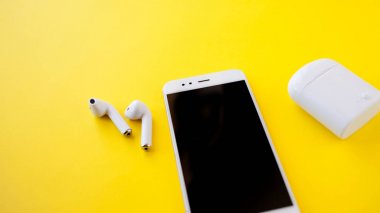 Beyaz kablosuz kulaklık ve akıllı telefon parlak sarı bir arka planda duruyor. Kulaklıklar şarj edilmiyor.