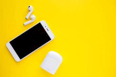 Beyaz kablosuz kulaklık ve akıllı telefon parlak sarı bir arka planda duruyor. Kulaklıklar şarj edilmiyor.