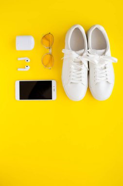 Beyaz spor ayakkabılar, pembe şeffaf güneş gözlükleri, kablosuz kulaklıklar ve akıllı telefonlar sarı arka planda yatıyorlar..