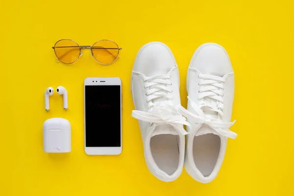 Witte sneackers, roze transparante zonnebrillen, draadloze koptelefoon en smartphone liggen op een gele achtergrond. — Stockfoto