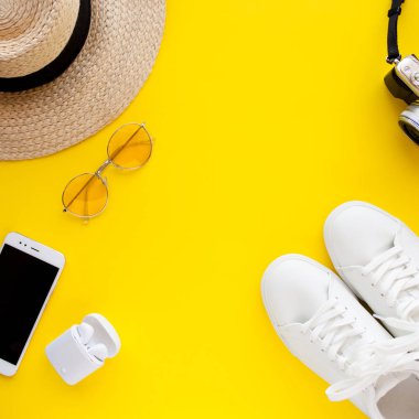 Beyaz spor ayakkabılar, şapka, güneş gözlüğü, kablosuz kulaklıklar, akıllı telefon ve fotoğraf makinesi sarı arka planda yatıyor.