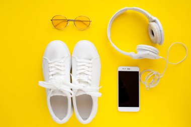 Beyaz spor ayakkabılar, pembe güneş gözlüğü, beyaz kulaklıklar ve akıllı telefon parlak sarı bir arka plan üzerinde yatıyor.