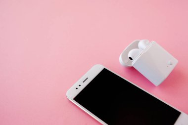 Beyaz kablosuz kulaklıklar ve akıllı telefon parlak pembe bir arka plan üzerinde yatıyor. Şarj durumunda kulaklıklar.