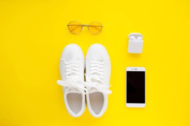 Beyaz spor ayakkabılar, pembe güneş gözlüğü, beyaz kablosuz kulaklıklar ve akıllı telefon parlak sarı bir arka plan üzerinde yatıyor.