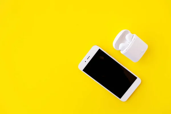 Witte draadloze hoofdtelefoon en smartphone liggen op een heldere gele achtergrond. Hoofdtelefoon in oplaadetui. — Stockfoto