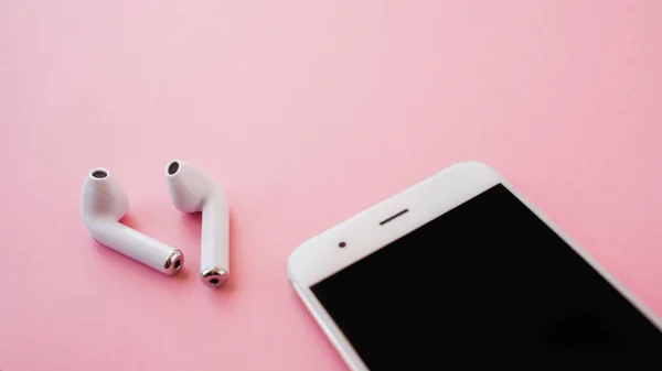 Los auriculares inalámbricos blancos y el teléfono inteligente se encuentran sobre un fondo rosa brillante. Auriculares fuera de la caja de carga . —  Fotos de Stock