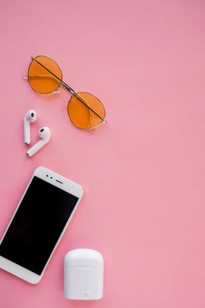 Las gafas de sol transparentes rosadas, los auriculares inalámbricos y el teléfono inteligente están sobre un fondo rosa . —  Fotos de Stock