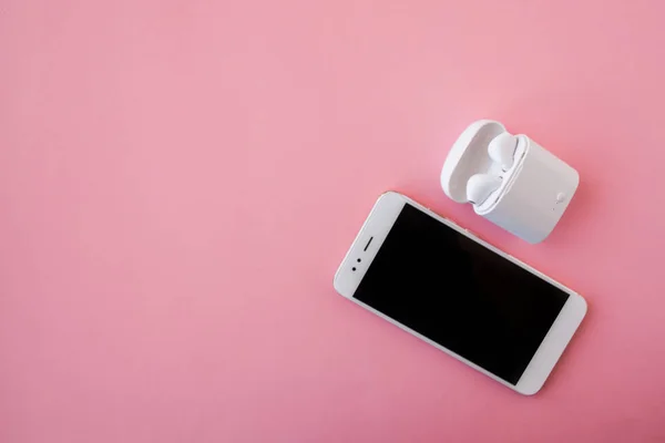 Los auriculares inalámbricos blancos y el teléfono inteligente se encuentran sobre un fondo rosa brillante. Auriculares en estuche de carga . —  Fotos de Stock